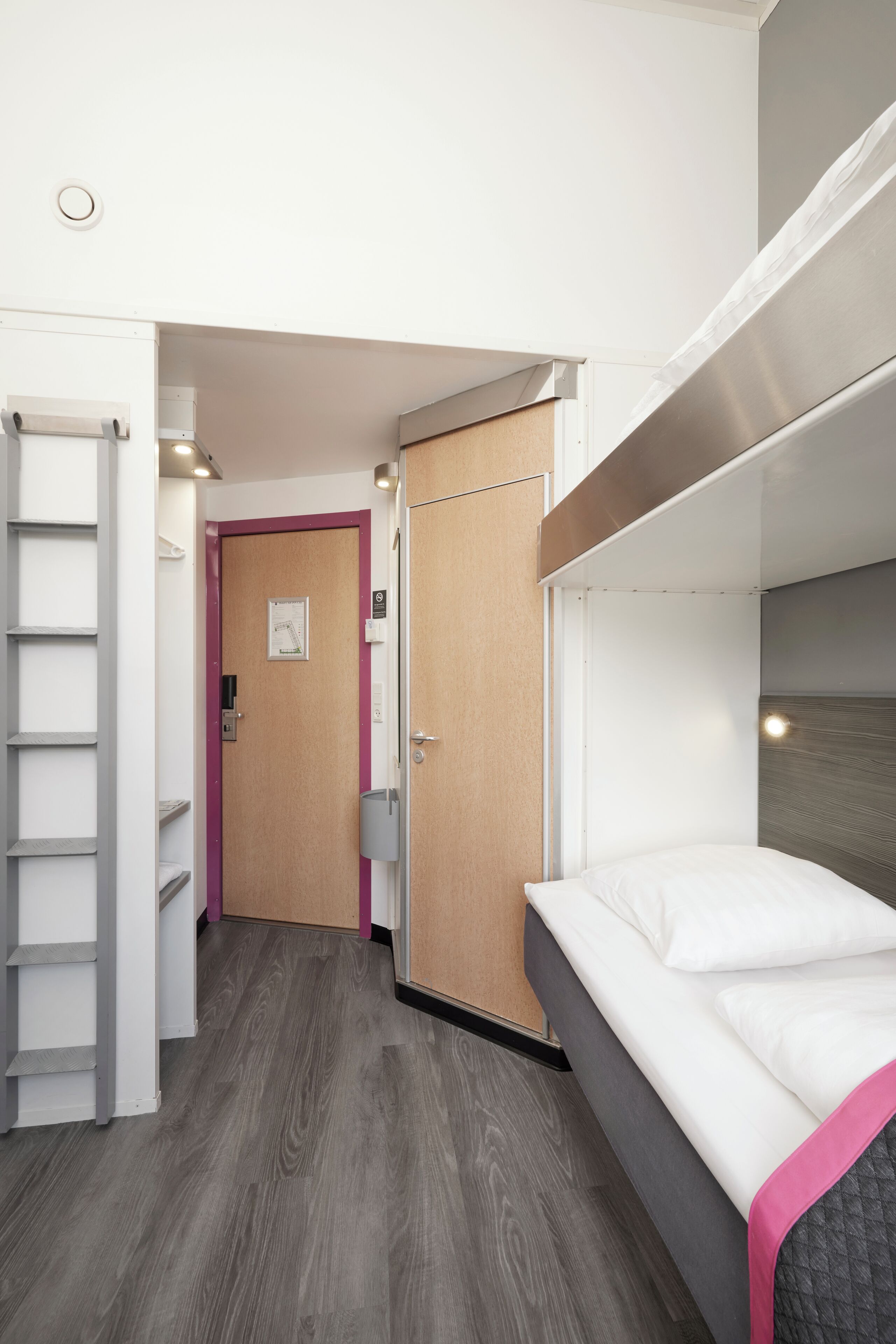 A compact hotel room with a bunk bed, a wooden door, a small closet, and a wooden floor.