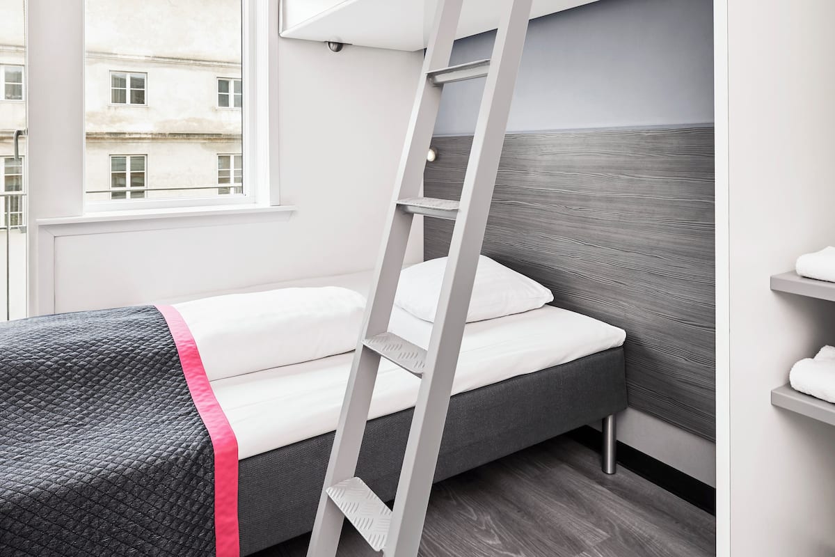 Une petite pièce avec un lit superposé, une échelle, une fenêtre et une vue sur les bâtiments environnants.