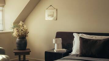 Un dormitorio con una cama grande, una mesita de noche, un jarrón con flores, un cuadro en la pared y una ventana.