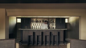 Una zona de bar con taburetes de madera, una barra con botellas y una persona en movimiento.
