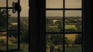 Vista a través de una ventana con un diseño de cuadrícula, mostrando un paisaje rural con árboles, césped y dos sillas de madera.