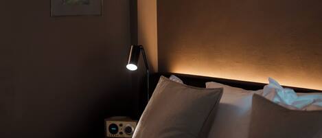 A dimly lit bedroom with a bed, pillows, a bedside table with a lamp, and a framed picture on the wall.