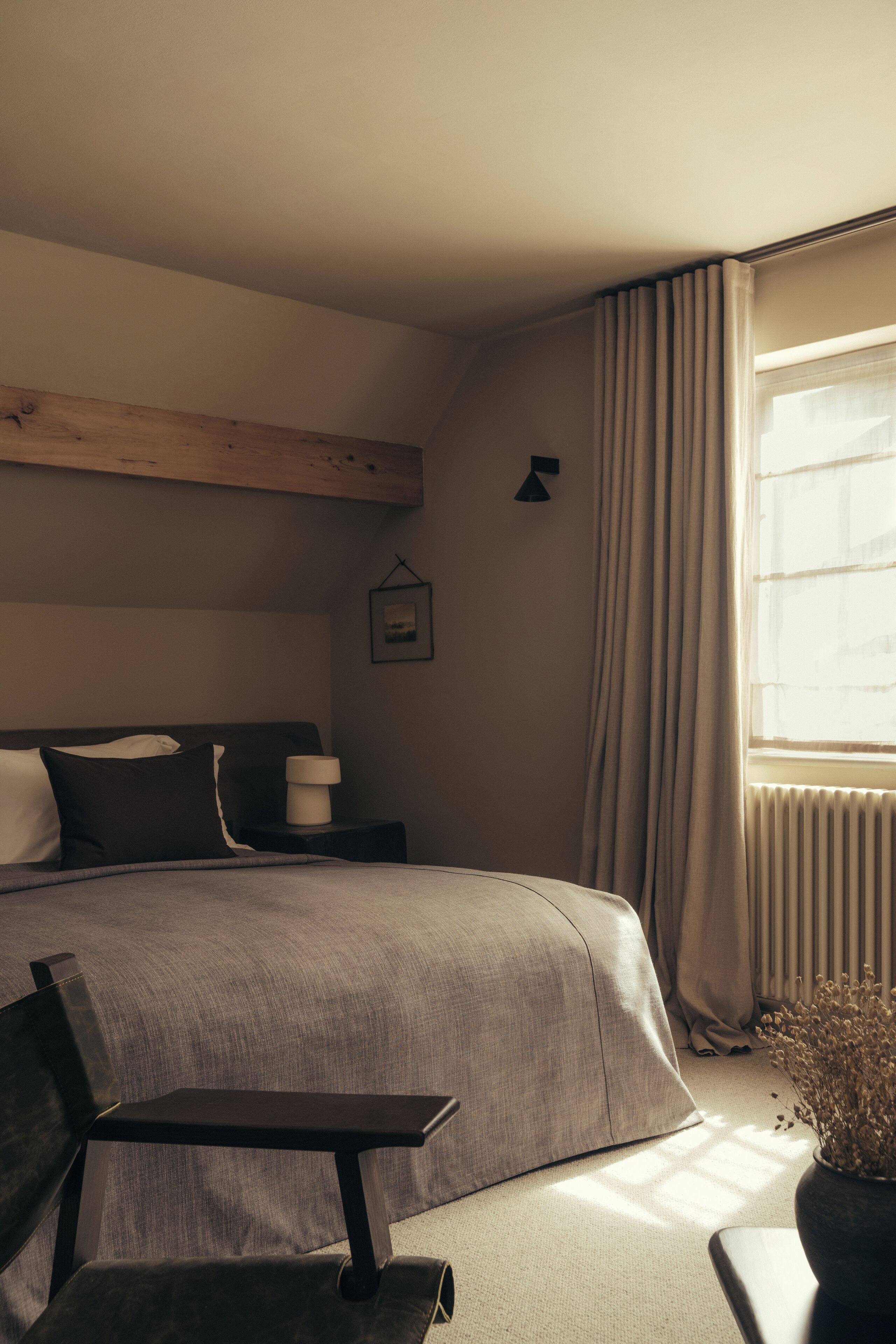 A bedroom with a large bed, a bedside table, a chair, a window with curtains, and a wall-mounted picture.