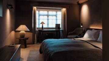 Habitación de hotel con cama, escritorio con lámpara, una silla, ventana con cortinas y radiador.