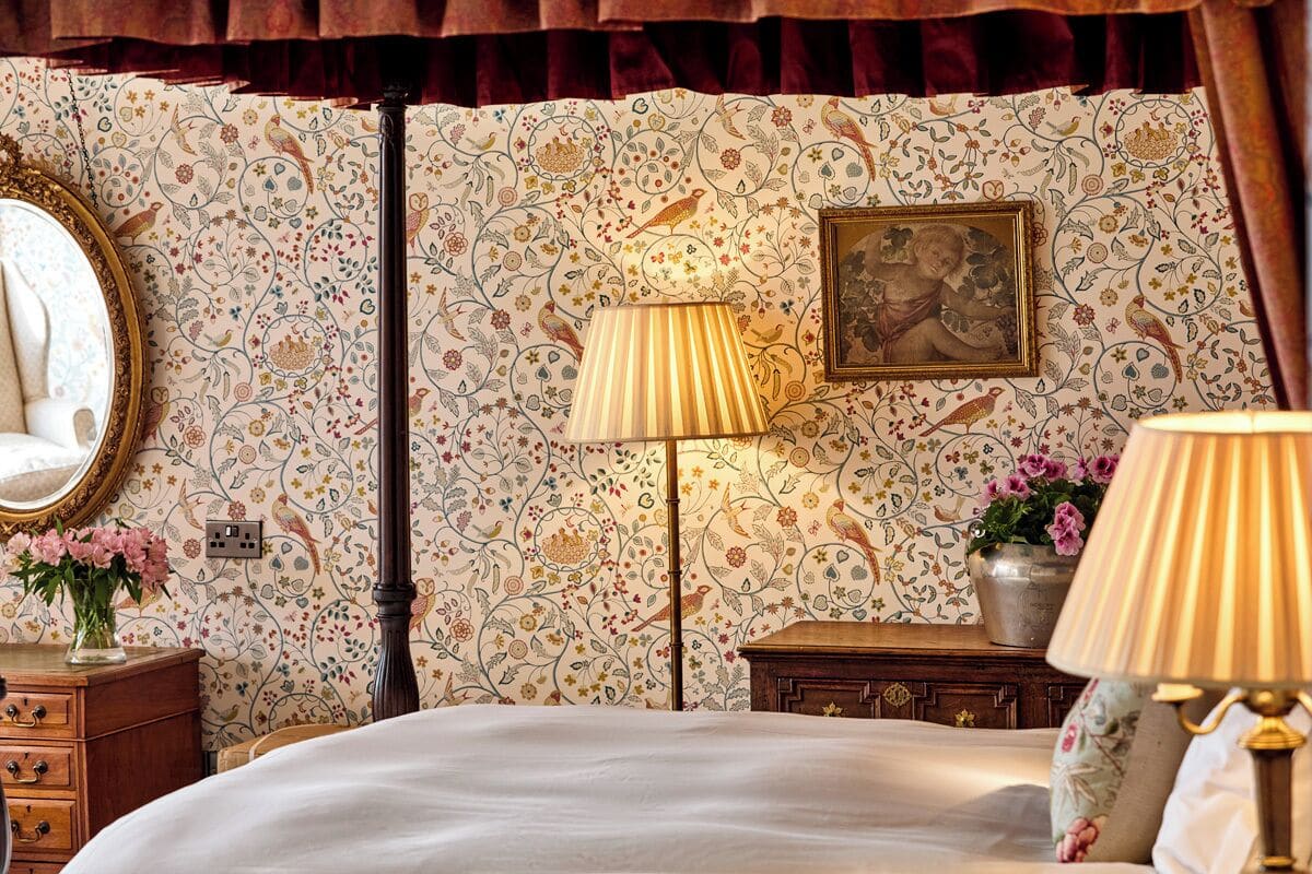 A bedroom with a bed, a nightstand, a lamp, a dresser, a mirror, and a floral wallpaper.