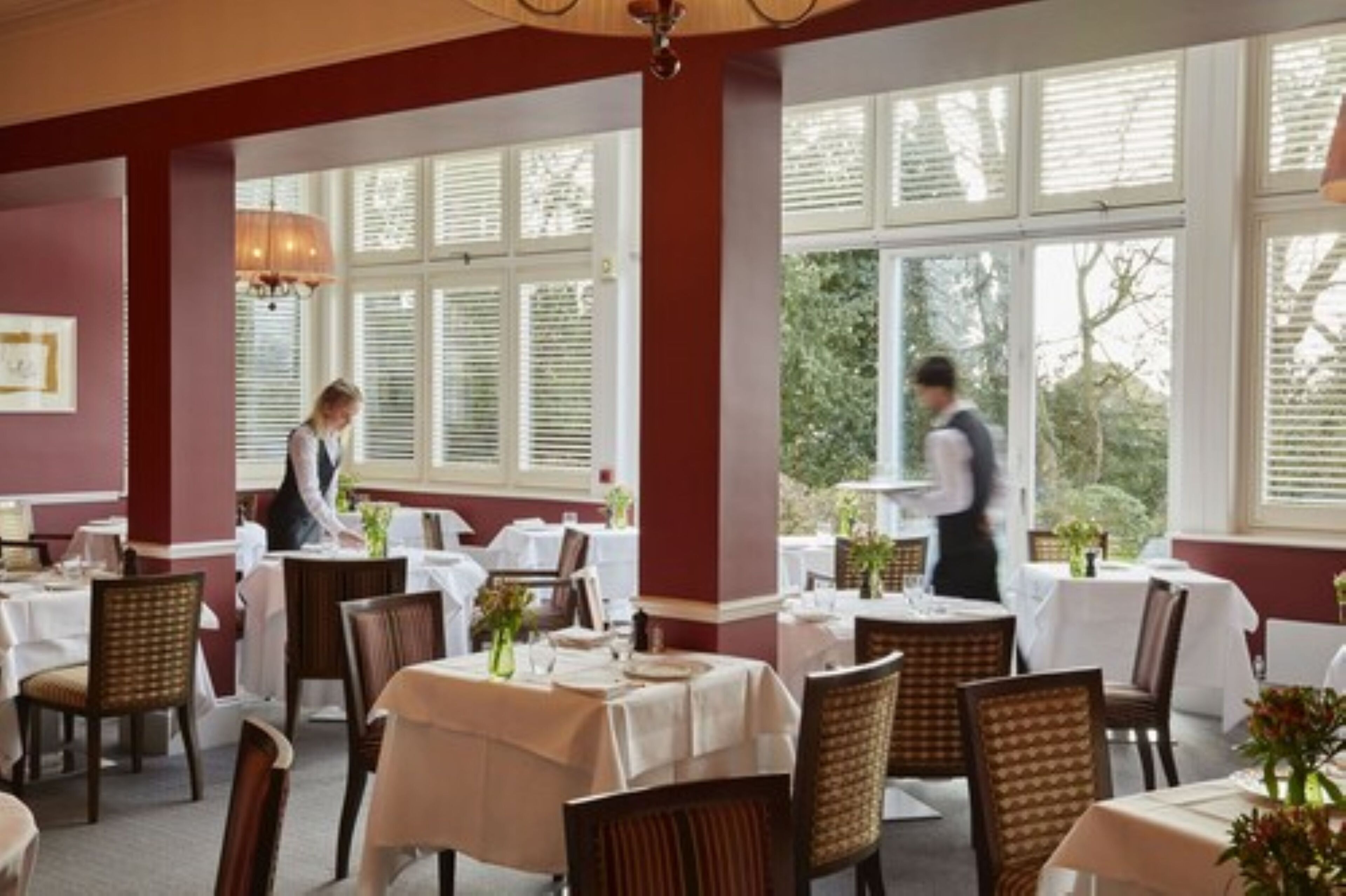 A restaurant with white tablecloths, wicker chairs, and large windows with blinds.