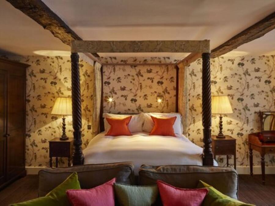 A four-poster bed with floral wallpaper, two bedside lamps, and a wooden nightstand with a lamp.