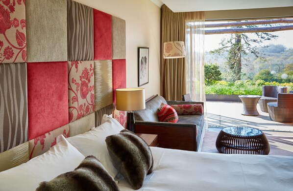Habitación de hotel con una cama grande, un sofá, una lámpara y vistas a un jardín y montañas.