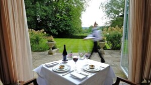 An outdoor dining area with a table set for two, surrounded by greenery and a view of the garden.