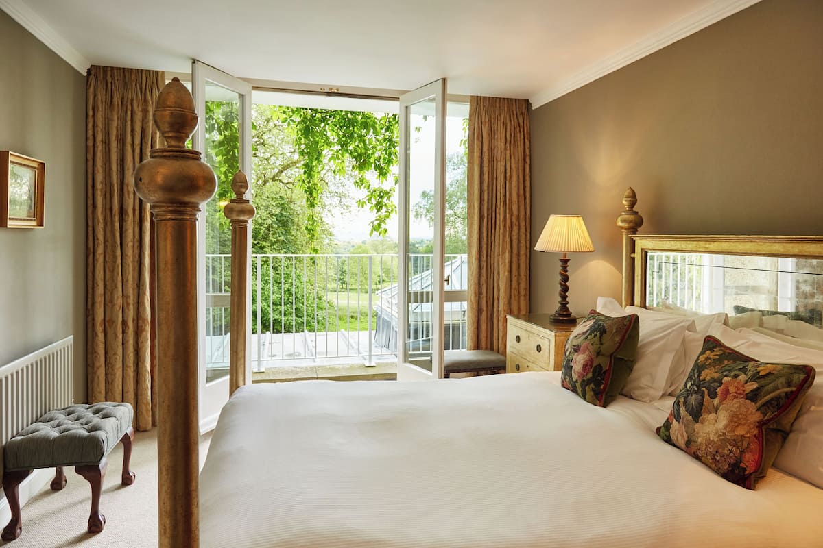A bedroom with a large bed, a nightstand, a lamp, a window with curtains, and a view of greenery outside.