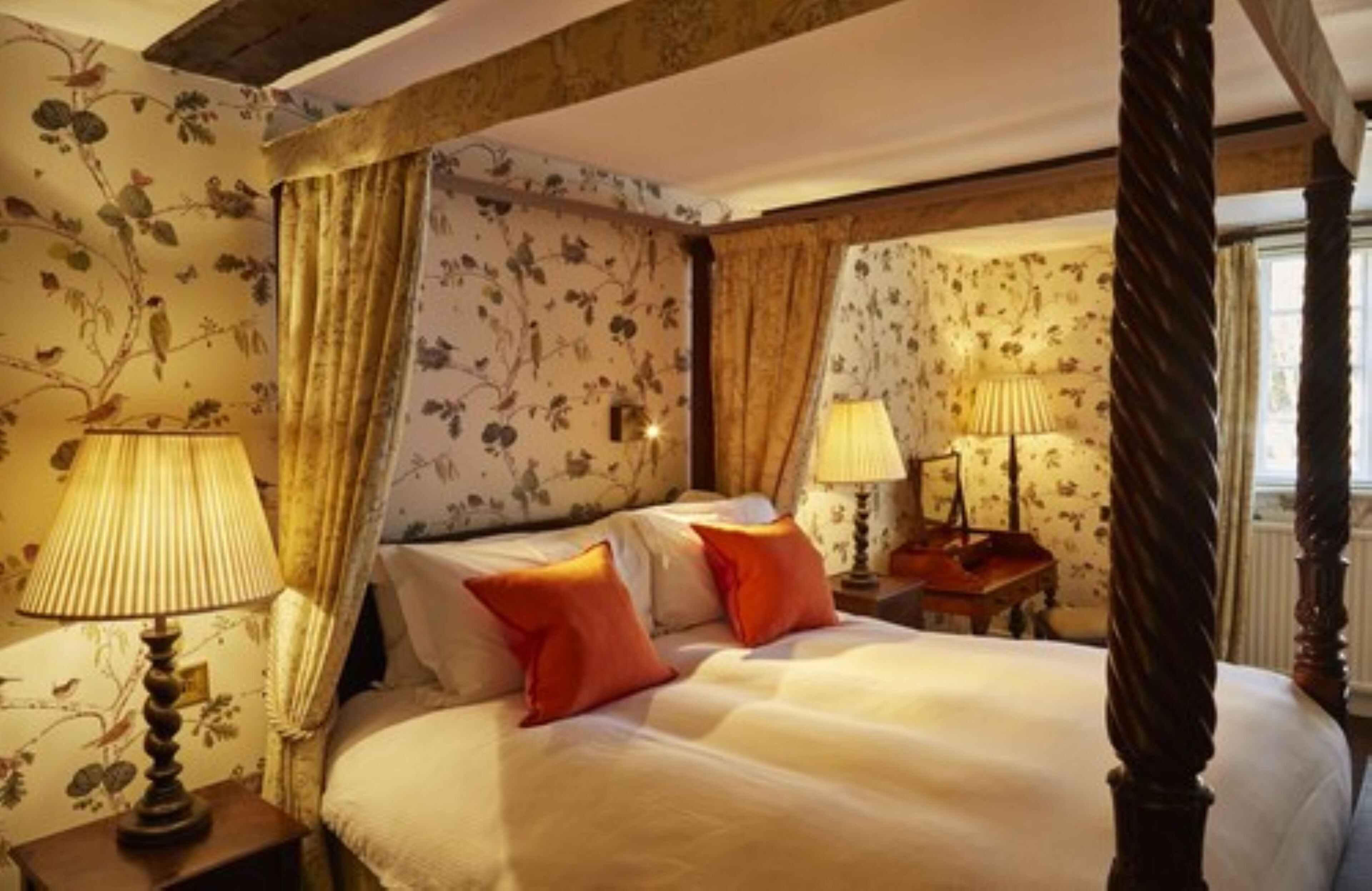 A bedroom with a four-poster bed, floral wallpaper, and two bedside lamps.