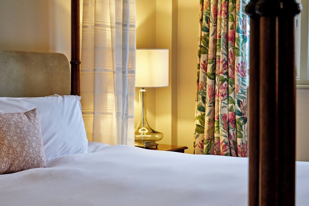 A neatly made bed with white linens and a floral pillow, a bedside lamp, and curtains with a floral pattern.