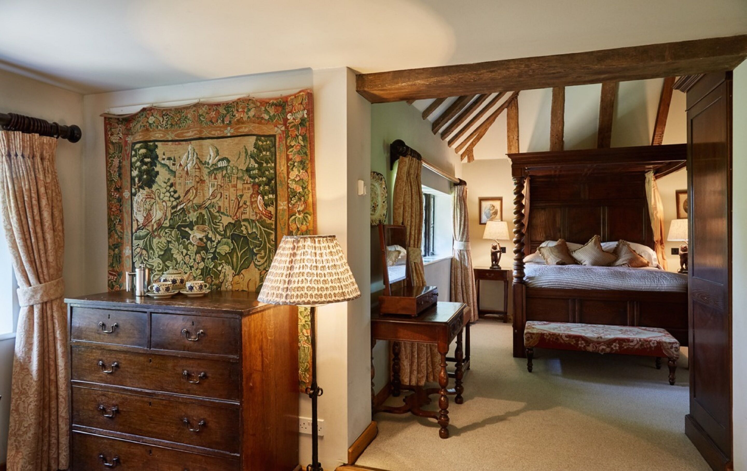 A traditional bedroom with a four-poster bed, a wooden dresser with multiple drawers, a tapestry on the wall, and a classic lamp.