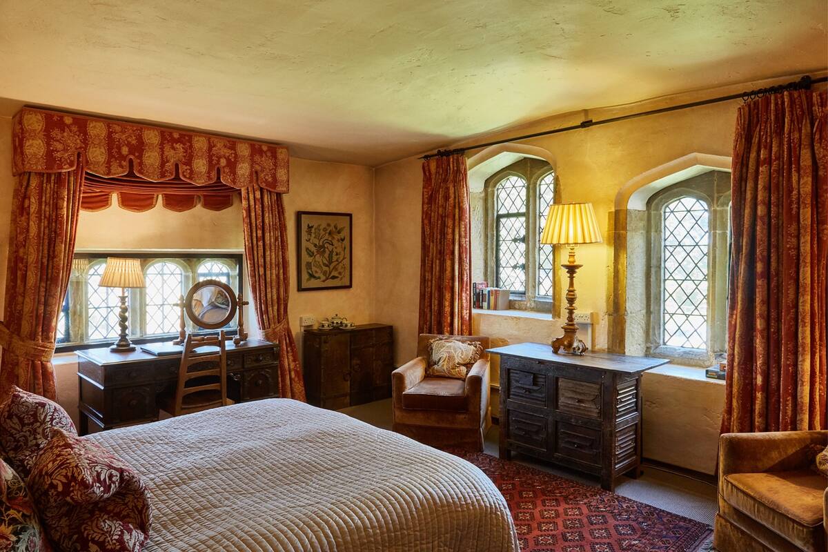 Une chambre avec un grand lit, un bureau, une chaise et une commode. Il y a des fenêtres avec des rideaux, une lampe et un tableau encadré au mur.