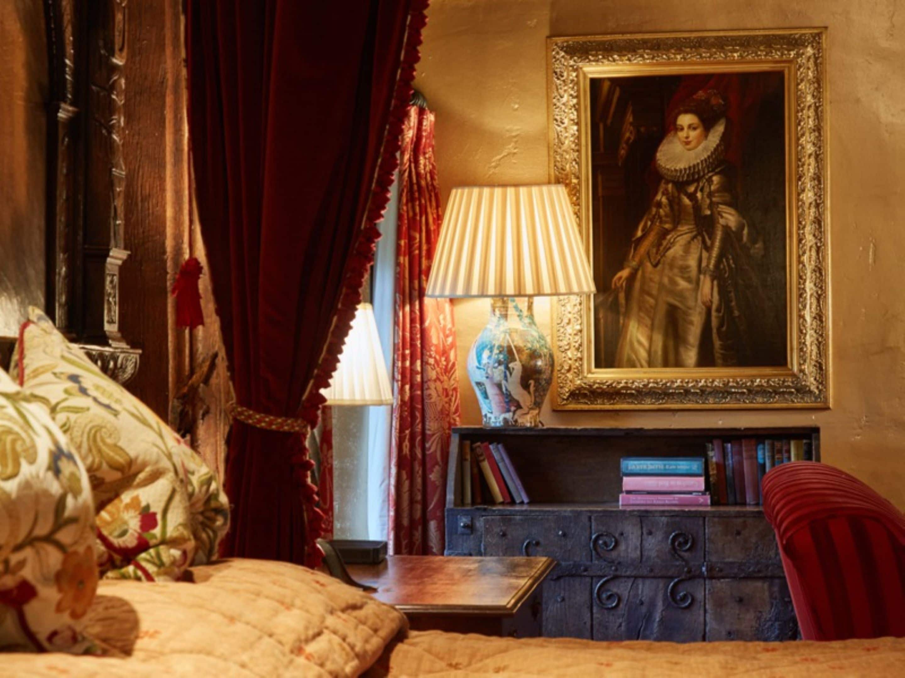 A bedroom with a bed, a nightstand with books, a lamp, a vase, and a framed portrait on the wall.