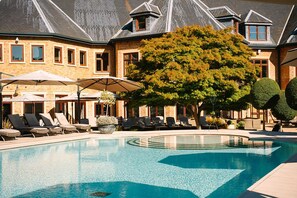 A swimming pool with lounge chairs and umbrellas, surrounded by a brick building with multiple windows and a tree. - Pennyhill Park (Bagshot)