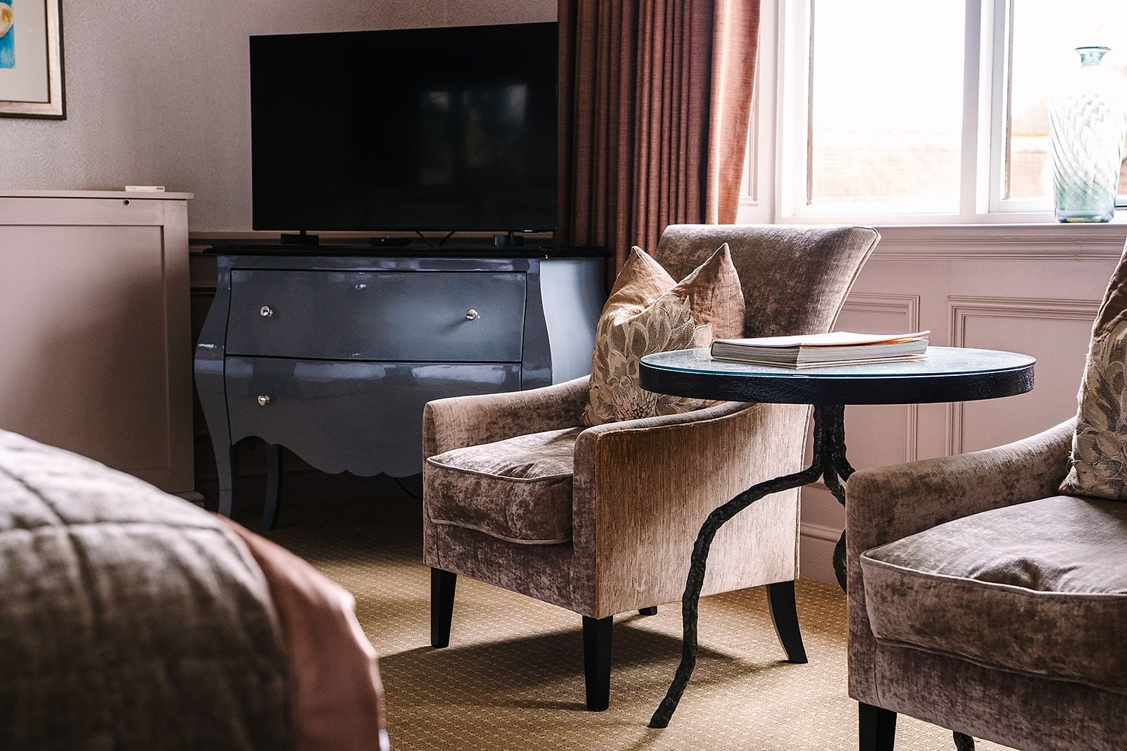A hotel room with a dark wooden dresser, a television, a round table with a book, and two armchairs.