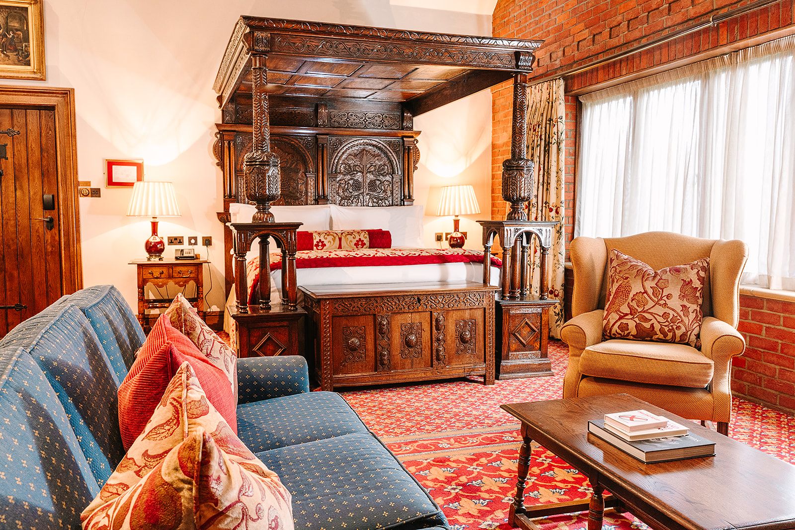A traditional room with a four-poster bed, a sofa, a chair, and a small table with books.