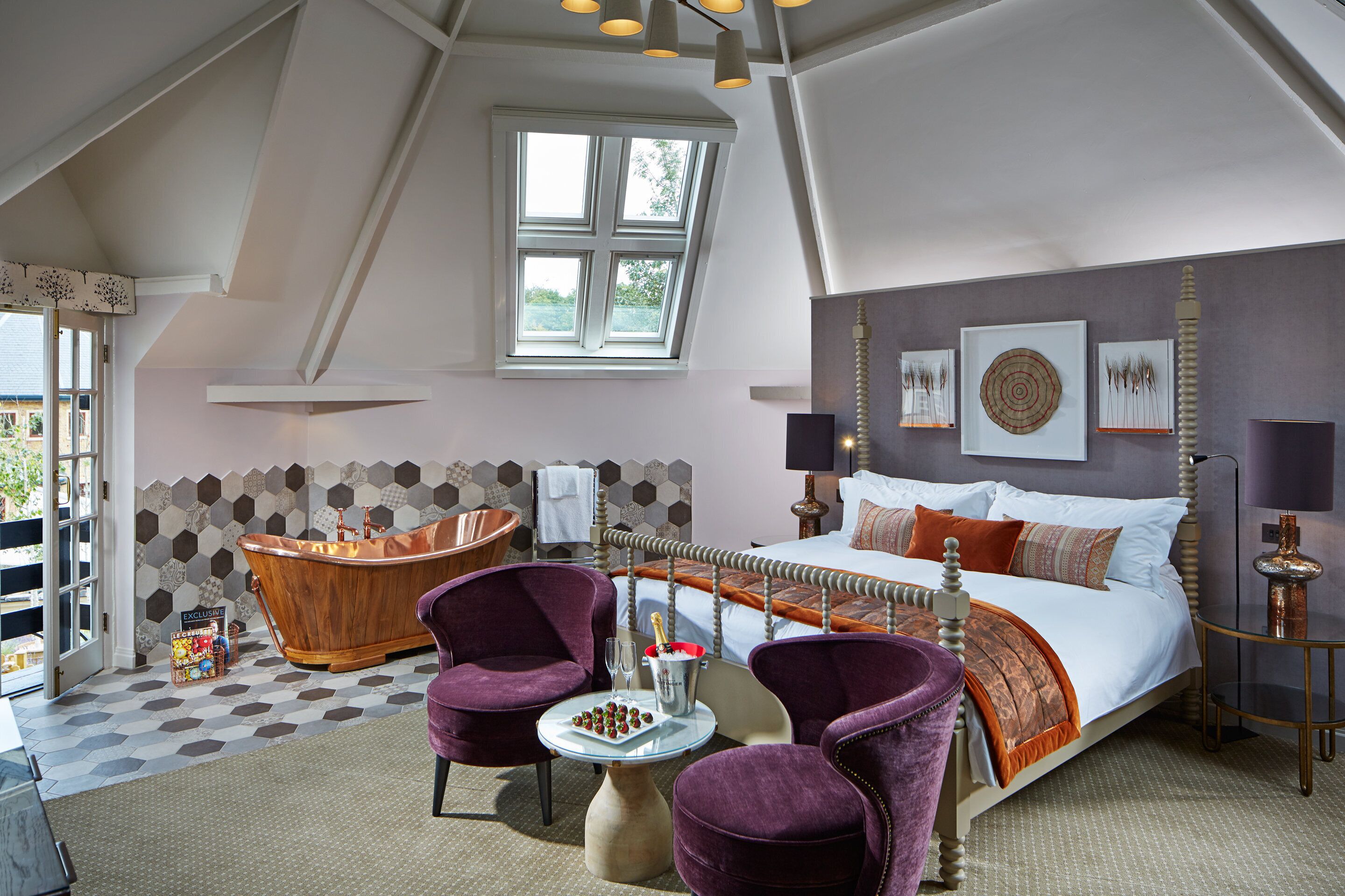 A bedroom with a large bed, a bathtub, two chairs, a table with a drink, and a view of the outdoors.