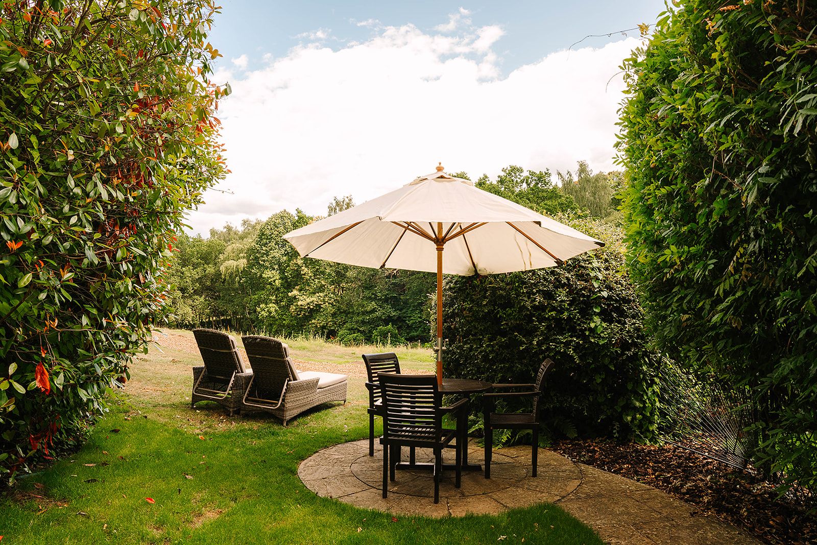 An outdoor patio with a table and chairs, surrounded by lush greenery and a large umbrella providing shade.