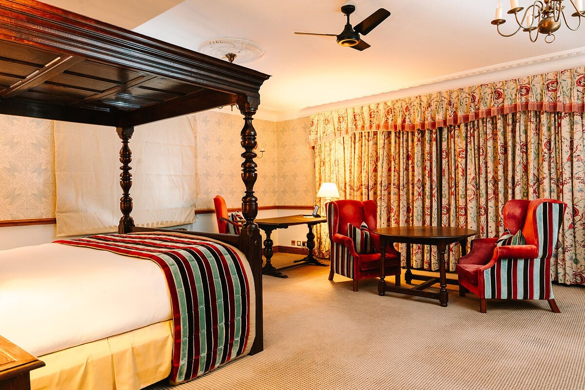 Habitación de hotel con cama con dosel, una mesa de comedor, dos sillones, una lámpara y un ventilador de techo.