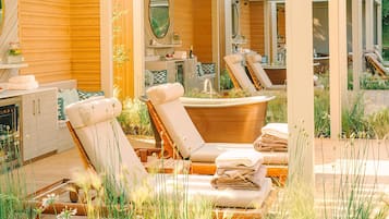 A spa area with two lounge chairs, a bathtub, and a wooden deck surrounded by plants.