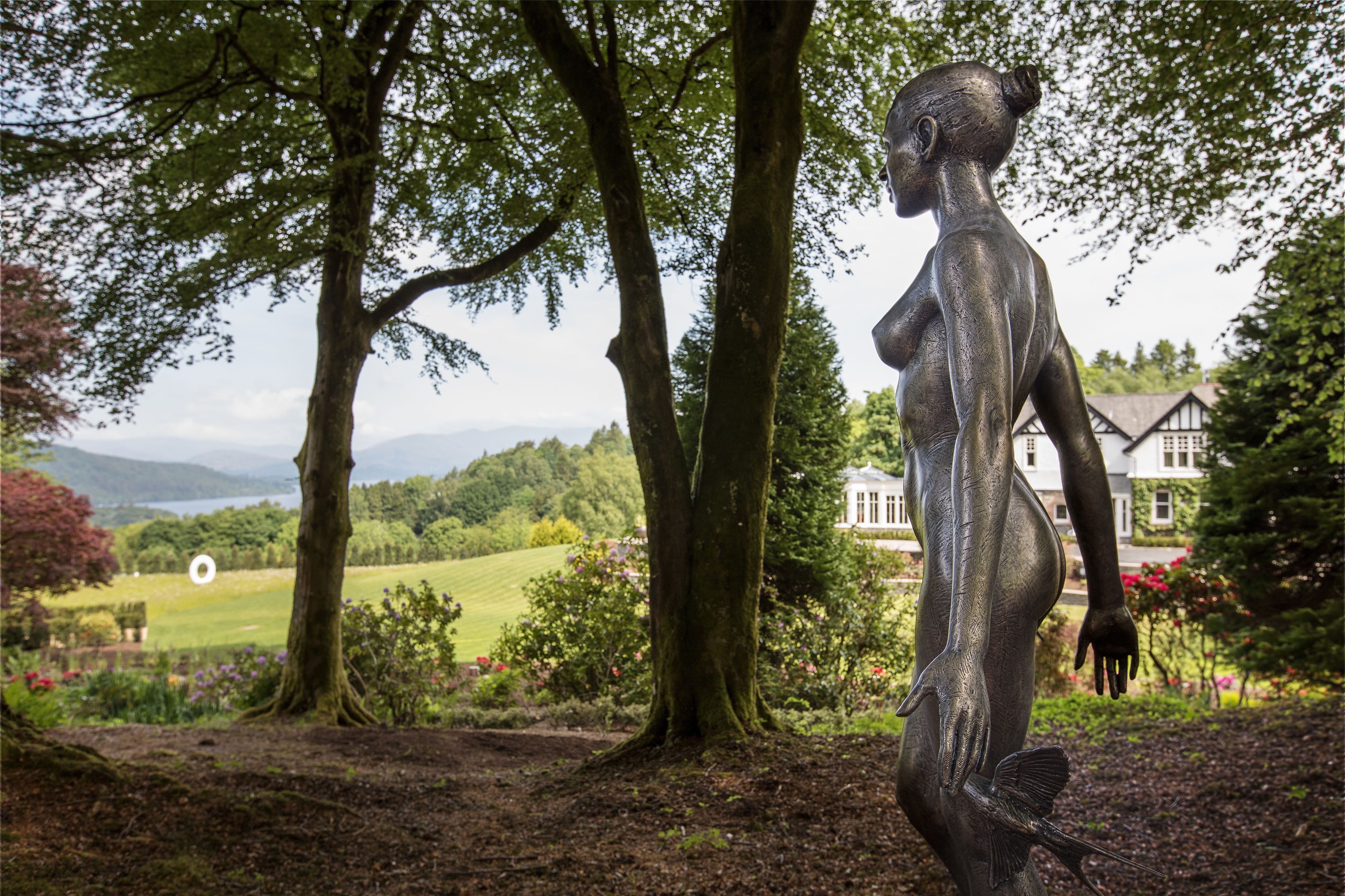 A statue of a woman in a garden with trees and a house in the background.