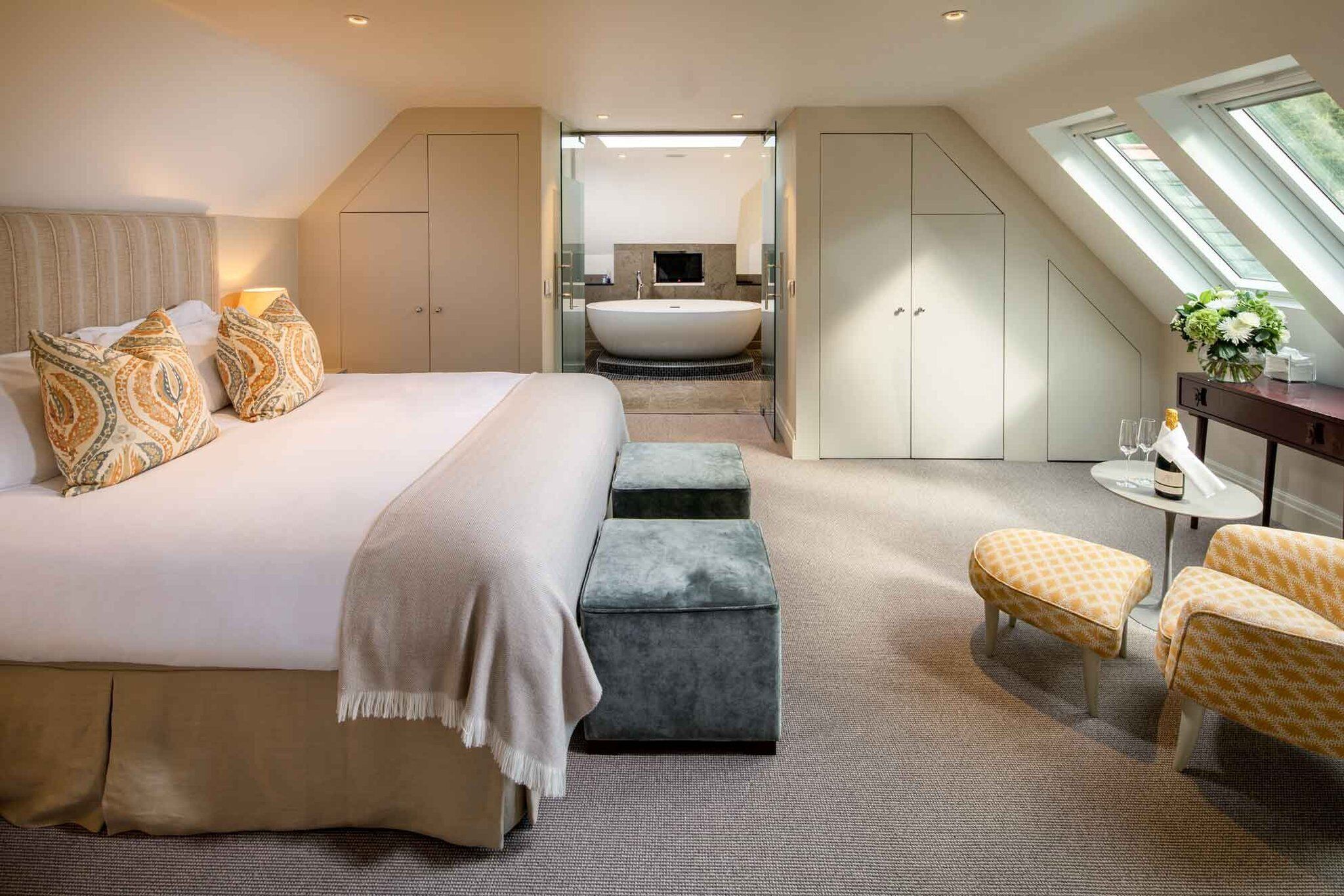 A modern hotel room with a large bed, a sitting area with ottomans, a small table with a vase, and a view of a bathroom with a bathtub.