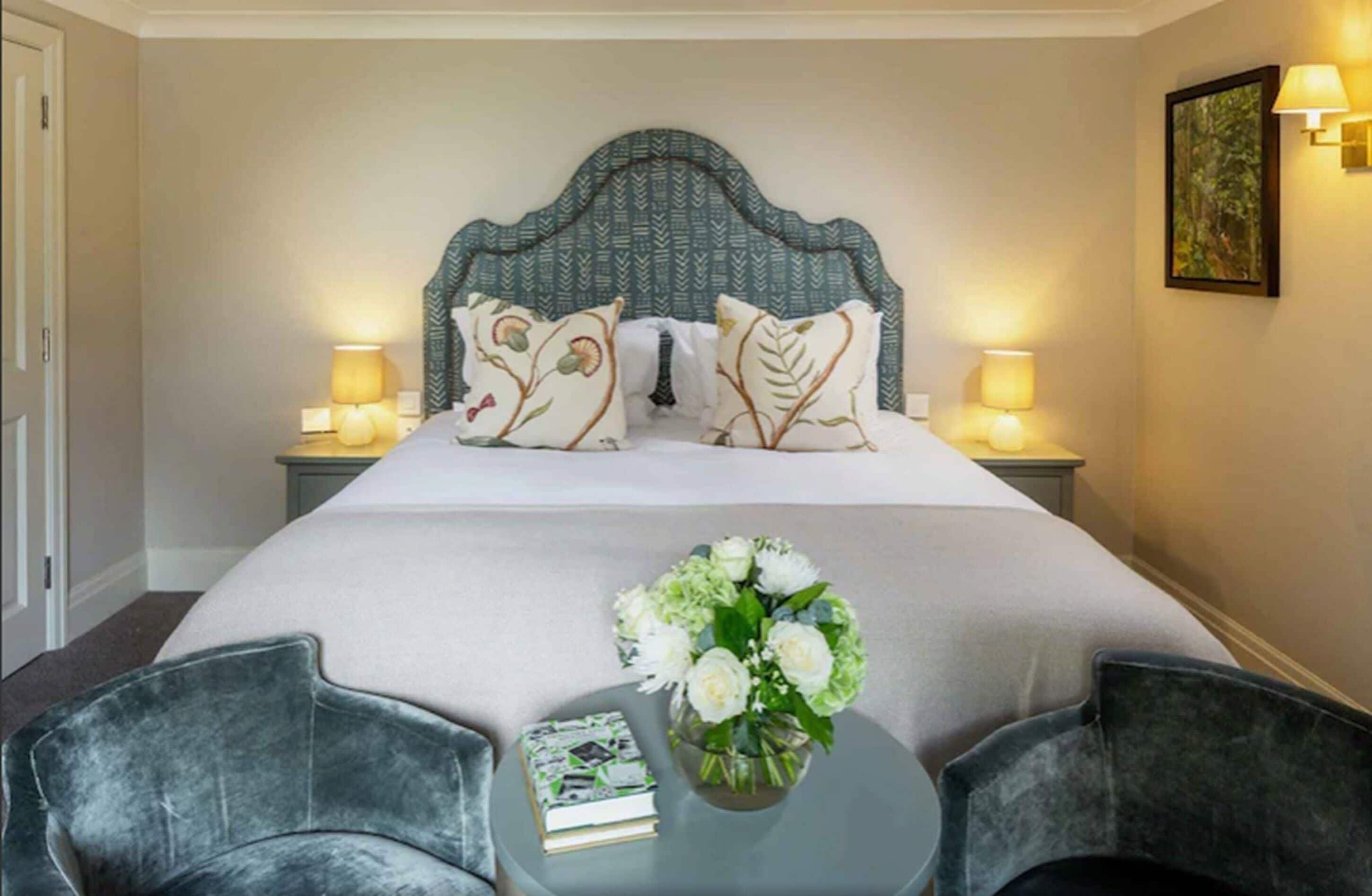 A hotel room with a large bed, two bedside lamps, a nightstand, a chair, a small table with a book and a vase of flowers.
