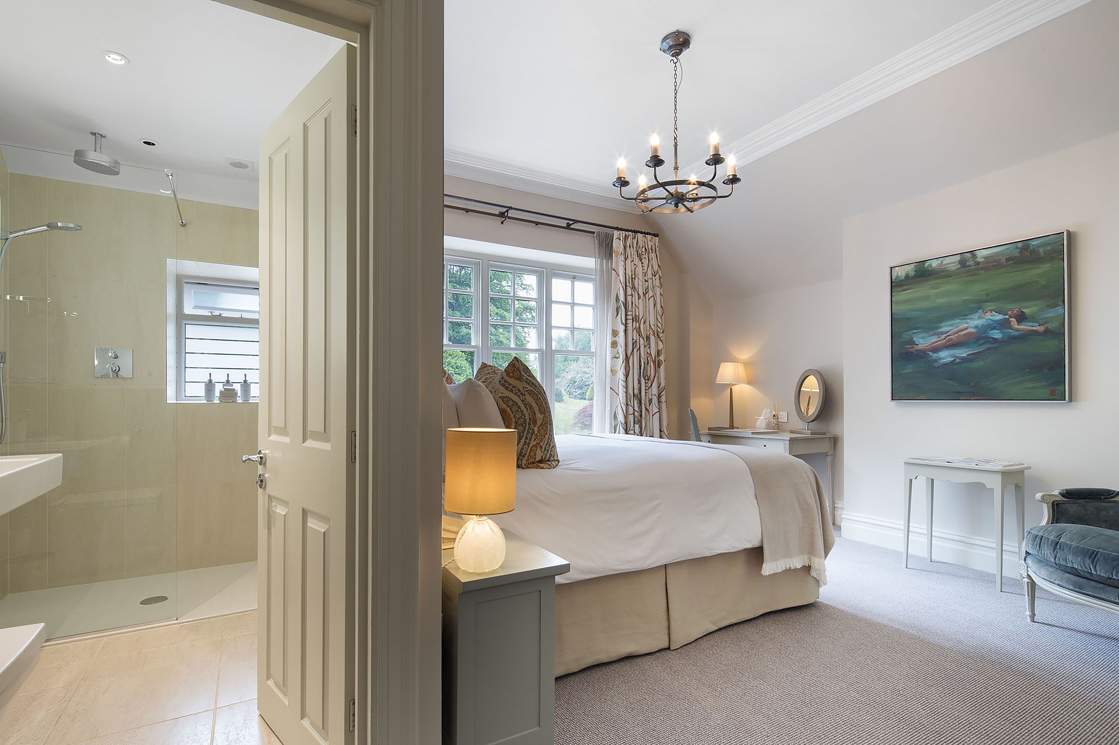 A bedroom with a large bed, a bedside table with a lamp, a painting on the wall, and a view of the garden through the window.