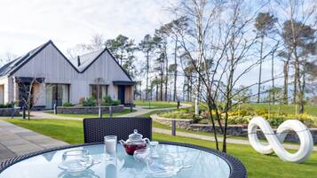 A glass-top table with tea set and cups, a wicker chair, and a modern building with a unique roof design in the background.