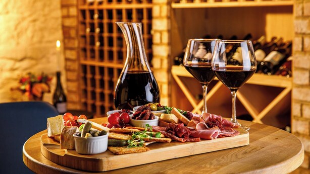Una tabla de fiambres con diferentes carnes, quesos y aceitunas sobre una tabla de madera, acompañada de dos copas de vino y una botella, todo sobre un fondo de estanterías llenas de vino.