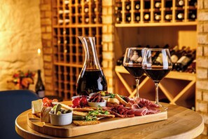A charcuterie board with various meats, cheeses, and olives on a wooden board, accompanied by two wine glasses and a bottle of wine, set against a backdrop of wine racks. - voco Lythe Hill Hotel & Spa by IHG (Haslemere)