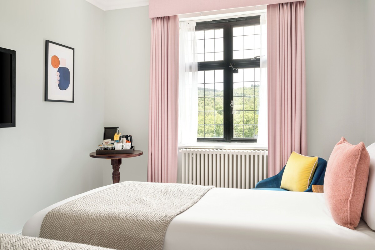 A bedroom with a bed, a bedside table with toiletries, a window with curtains, and a framed picture on the wall.