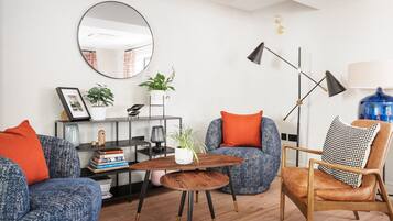 Un salon moderne avec un grand miroir rond, une table basse en bois et deux canapés bleus à motifs, agrémentés de coussins orange.