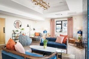 A modern living room with a blue sofa, a round coffee table, and a blue armchair. There is a bookshelf with books and a plant, and a window with patterned curtains. - voco Lythe Hill Hotel & Spa by IHG (Haslemere)