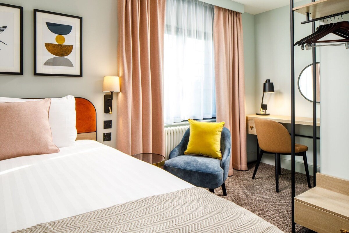 A hotel room with a bed, a desk, a chair, a lamp, and a wardrobe.