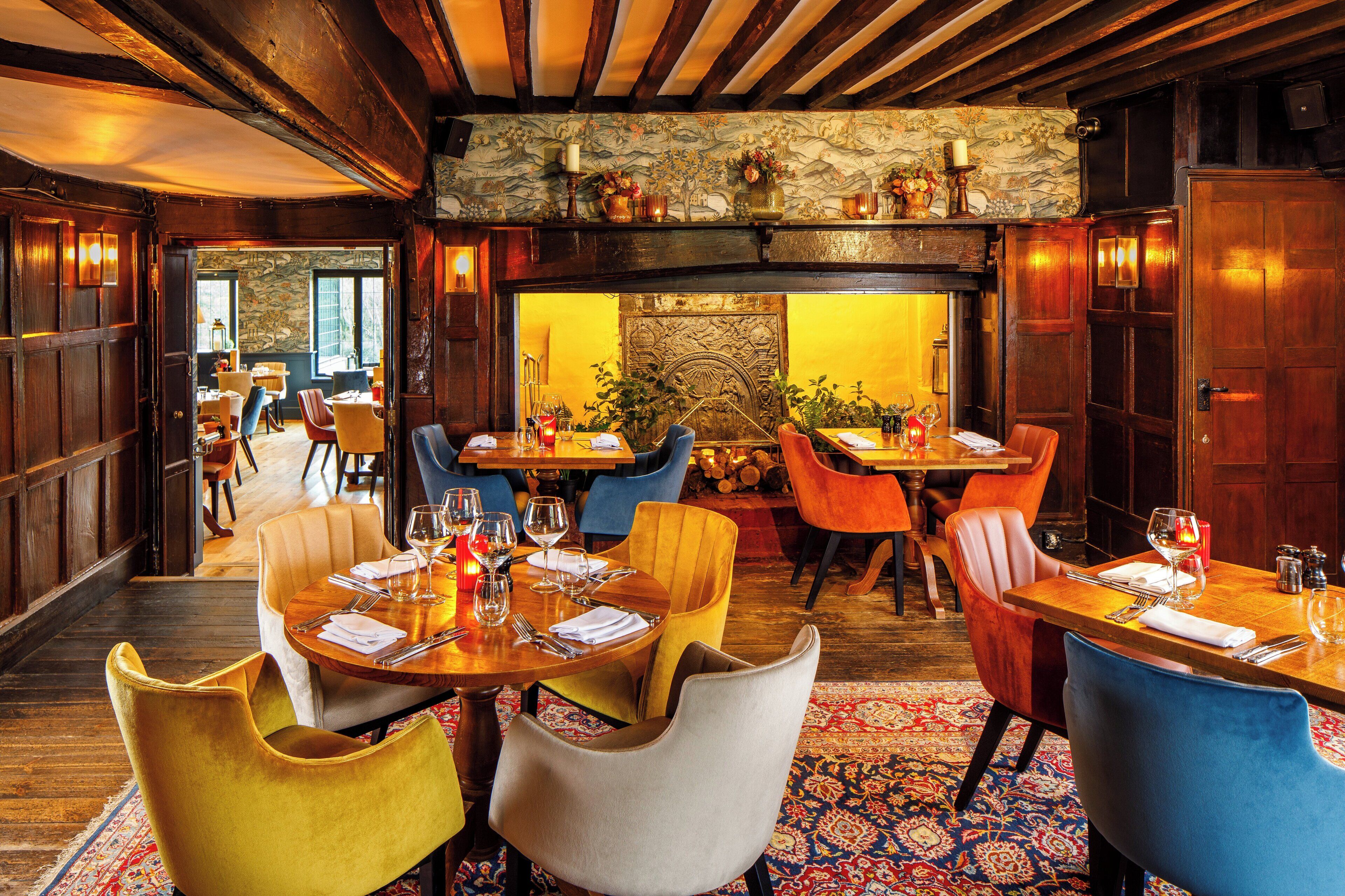 Un restaurante con vigas de madera, una pared de piedra y una alfombra estampada. Las mesas están preparadas con sillas y copas de vino.