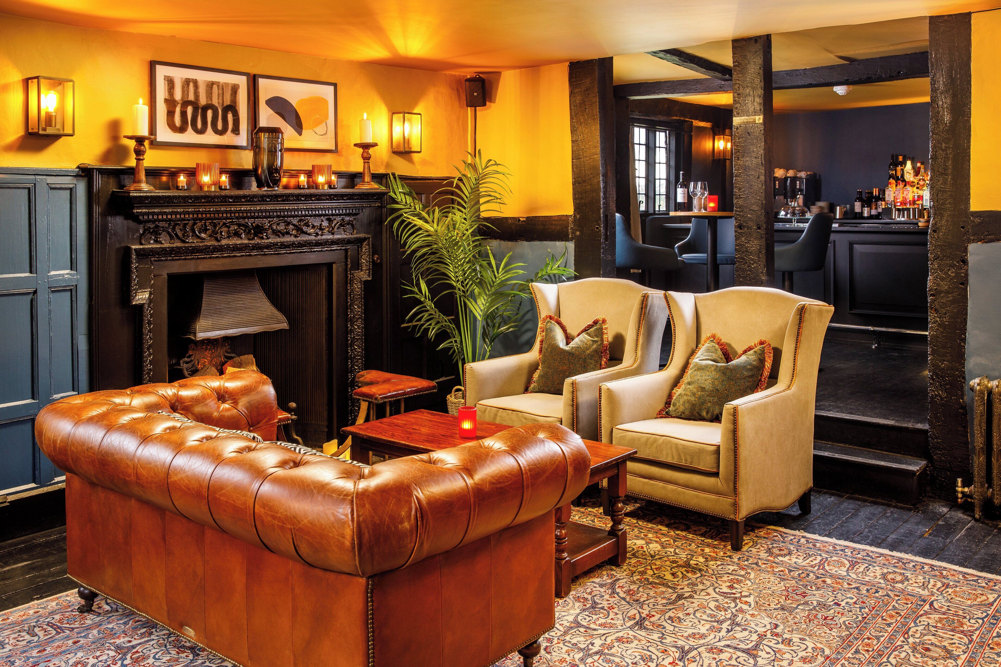 A cozy lounge area with a leather sofa, armchairs, a fireplace, and a bar in the background.