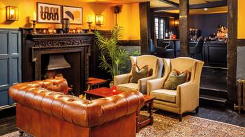 A cozy lounge area with a leather sofa, armchairs, a fireplace, and a bar in the background.