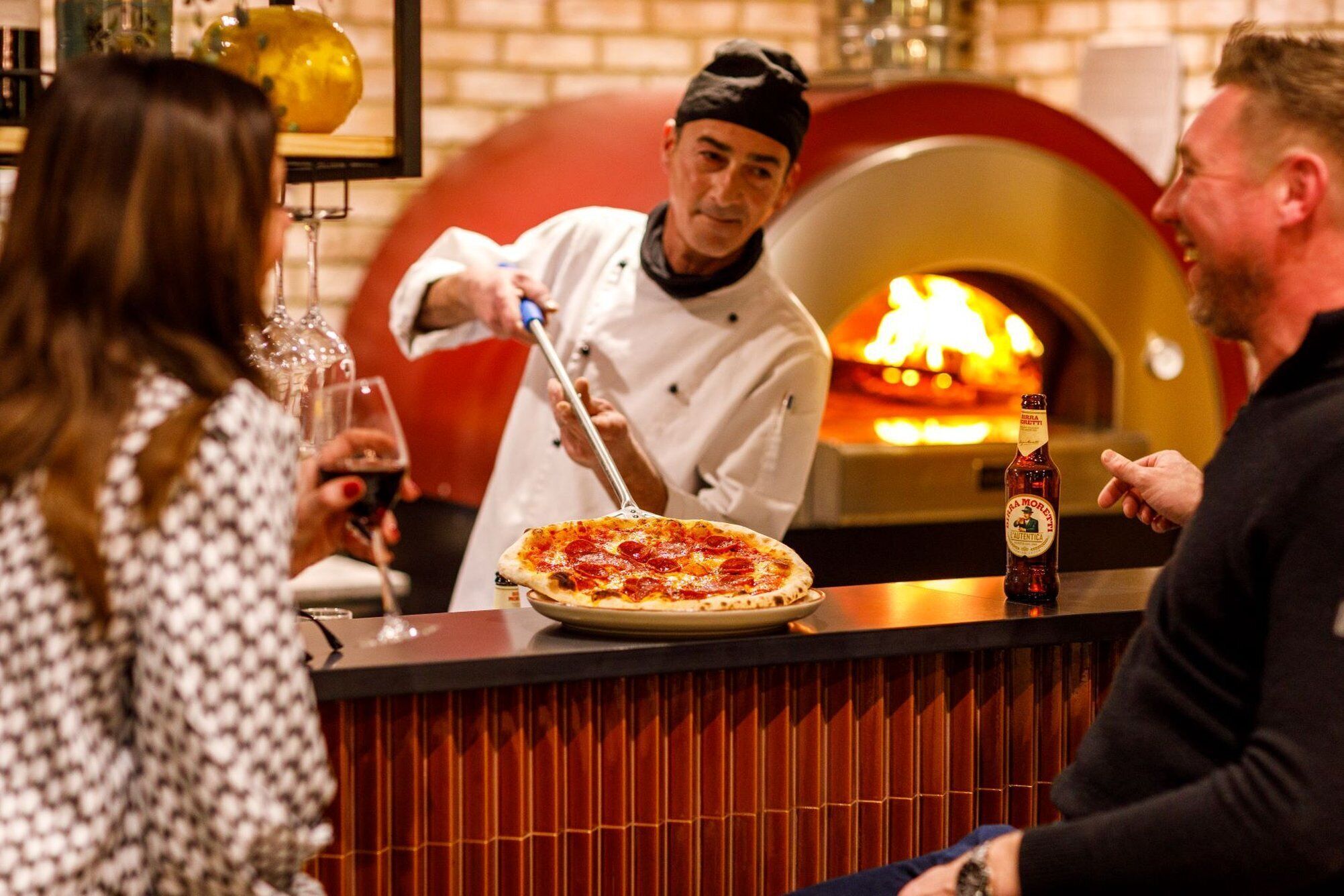 A pizza being served at a restaurant with a brick oven in the background.