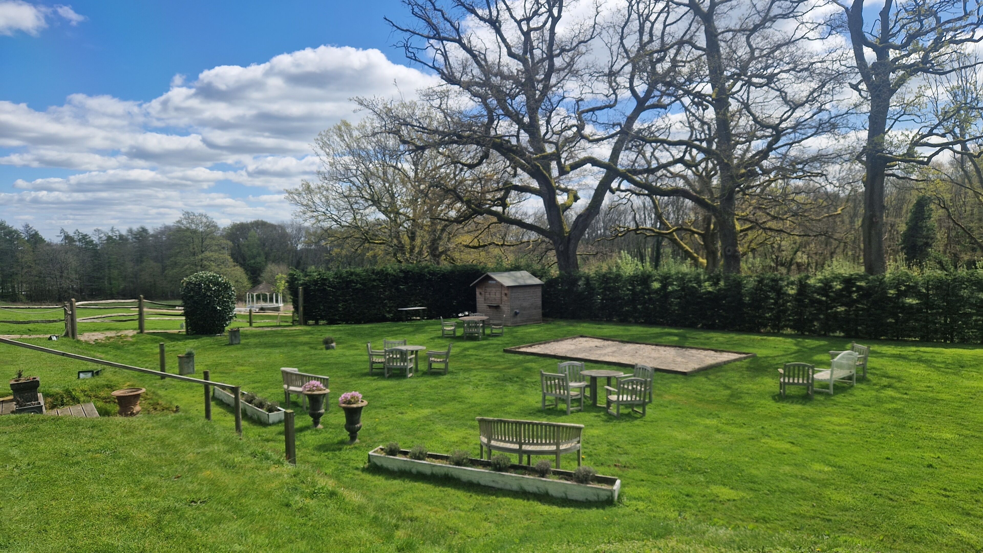 A spacious outdoor area with a grassy lawn, several wooden benches, a small wooden structure, and a large tree.