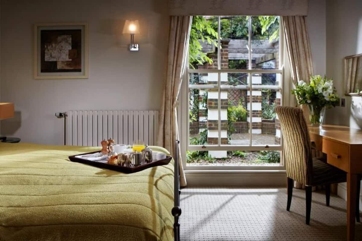A hotel room with a bed, a desk, a chair, a radiator, a window with curtains, and a tray with breakfast items.