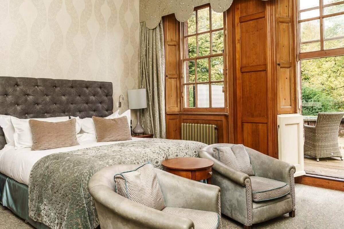 A bedroom with a large bed, two armchairs, a small table, and a view of the outdoors through large windows.
