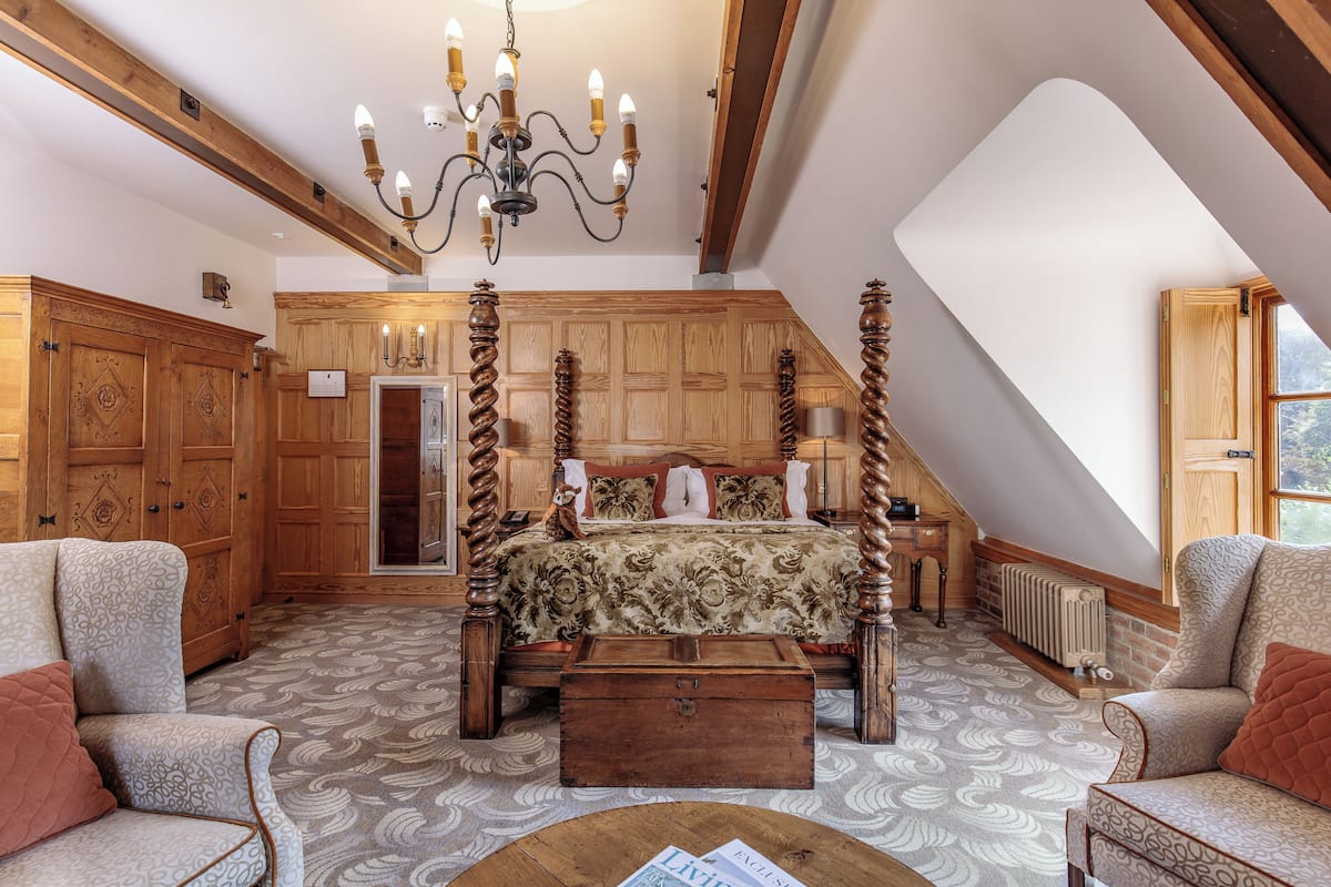 A four-poster bed with a floral bedspread, wooden paneling on the walls, a chandelier, and a view of the outdoors through a window.