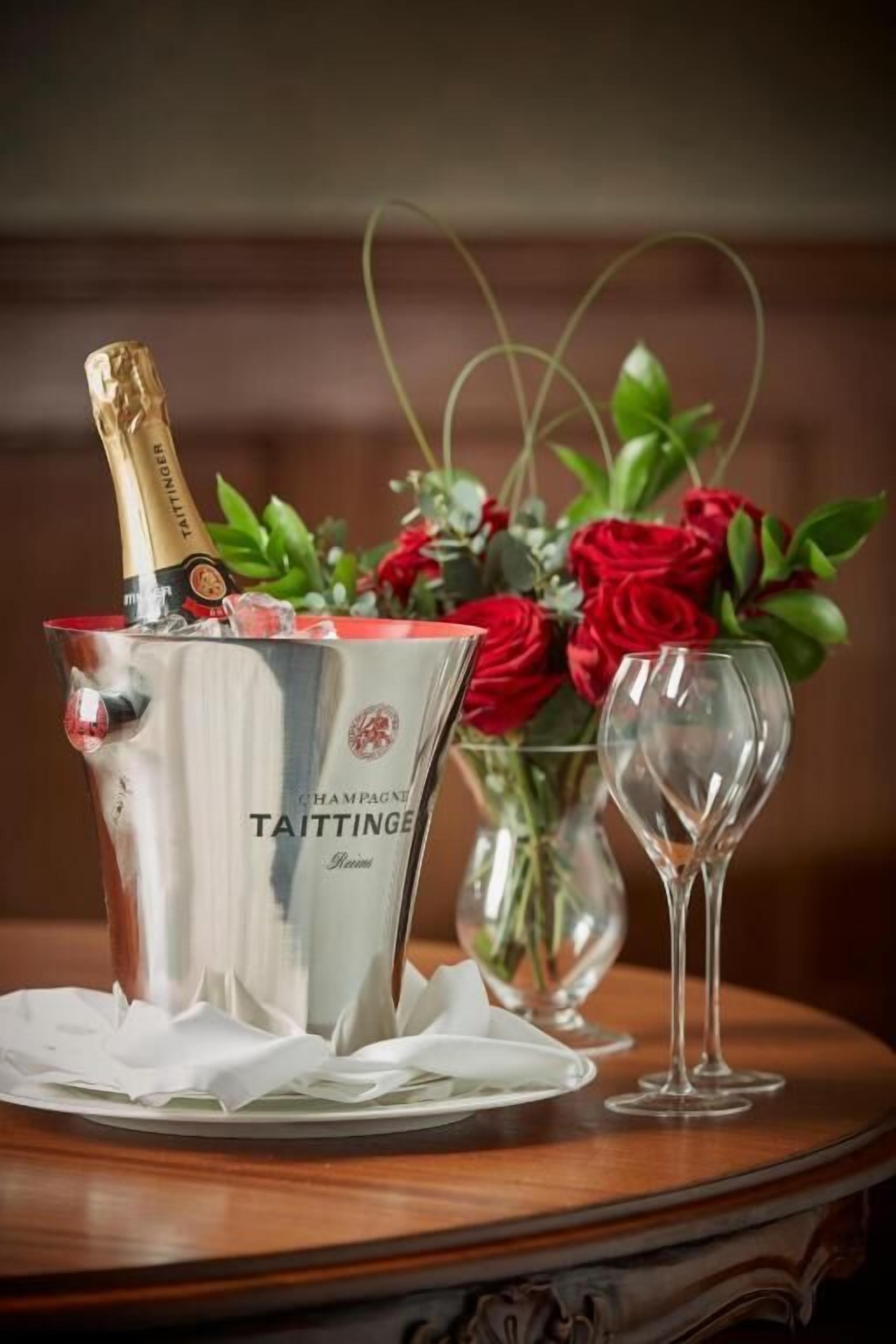 A bottle of champagne in a silver bucket, a glass of champagne, and a vase with red roses on a wooden table.