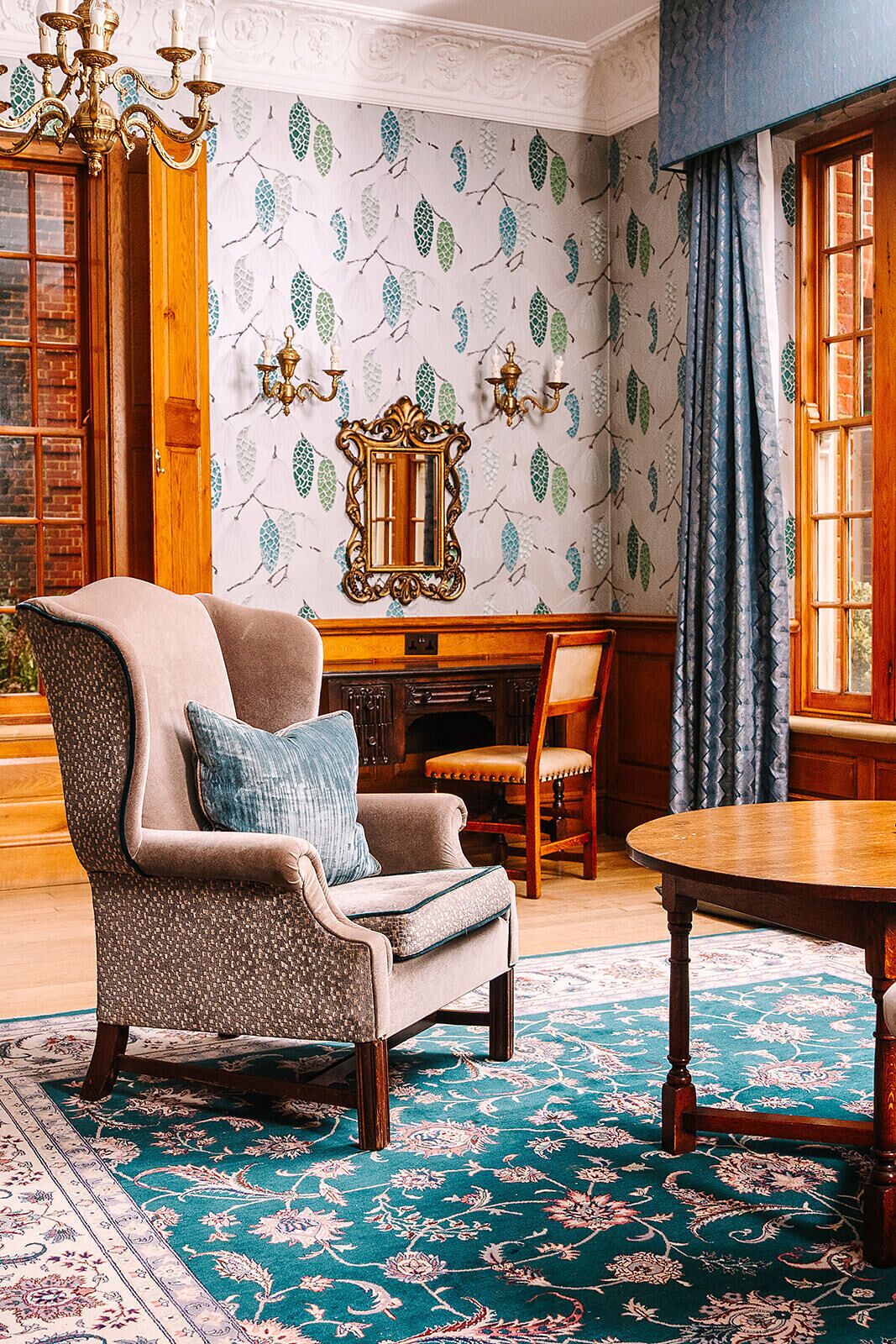 Une pièce avec un papier peint à motifs, un fauteuil classique agrémenté d’un coussin, une table en bois et un tapis à motifs.
