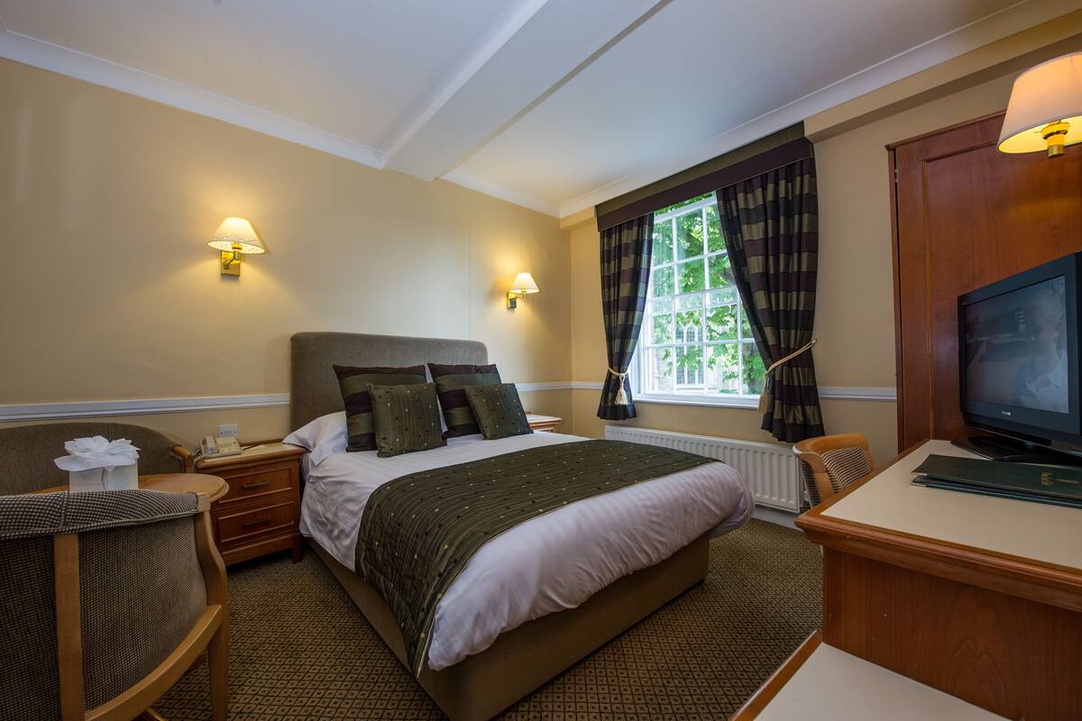 A hotel room with a bed, a desk, a chair, a television, and two wall-mounted lamps.