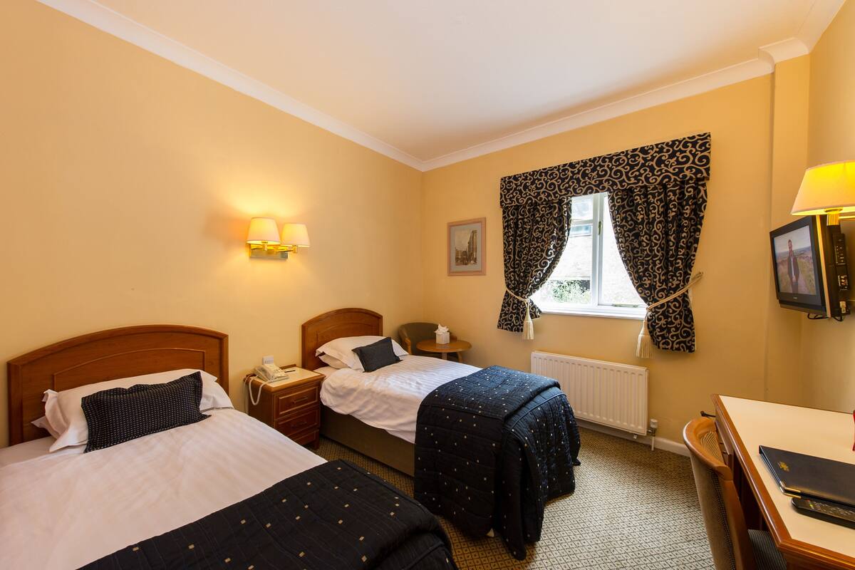A hotel room with two beds, a desk, a chair, a lamp, a window with patterned curtains, and a picture on the wall.