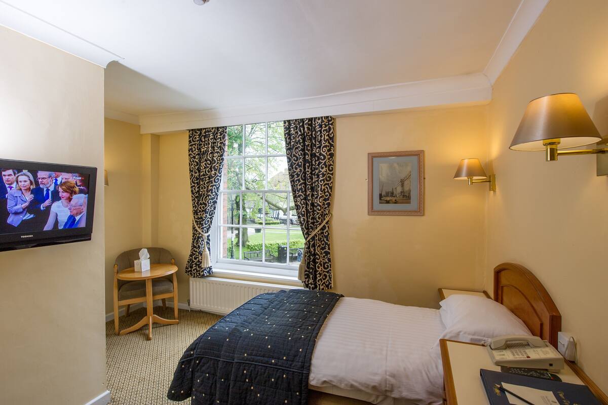 A hotel room with a bed, a bedside table with a phone, a lamp, a TV on the wall, a chair, and a window with curtains.